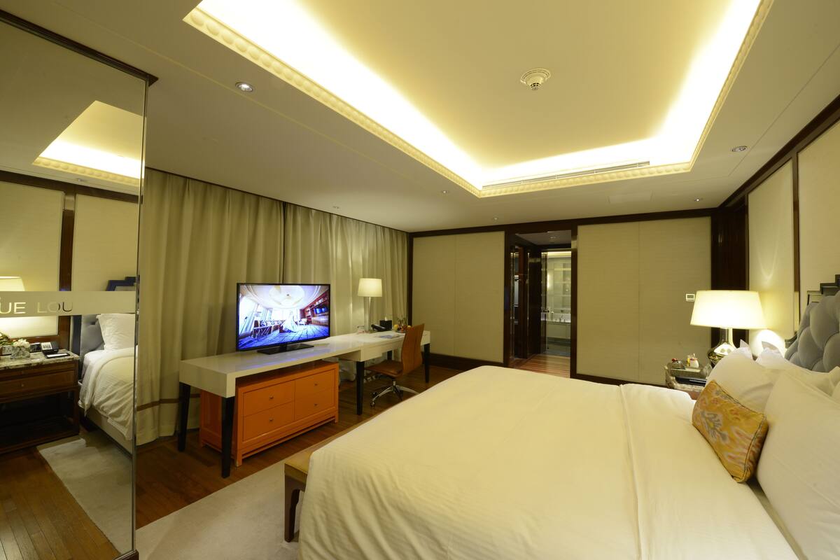 Habitación de hotel con una cama grande, un escritorio con televisor, una silla y una mesita de noche con lámpara.