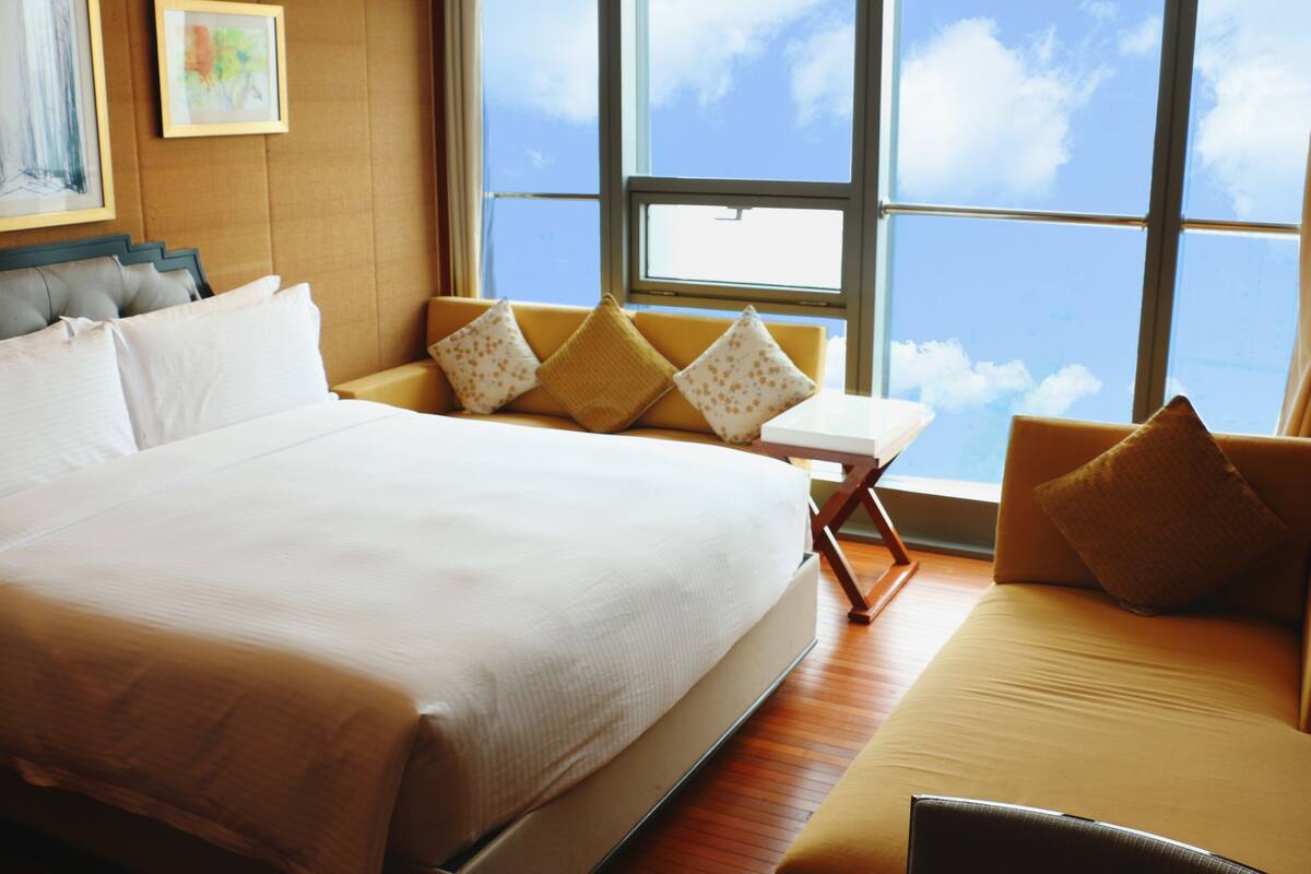 Una cama grande con sábanas blancas, un sofá con cojines amarillos y una silla con un cojín amarillo en una habitación con un ventanal que ofrece vistas al cielo.