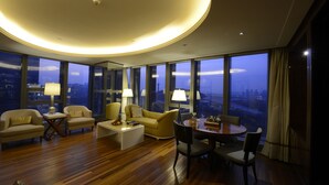 Una habitación de hotel moderna con una lámpara de techo circular, ventanas amplias que ofrecen vistas a la ciudad, un sofá amarillo, una mesa de comedor con sillas y una mesa de centro.