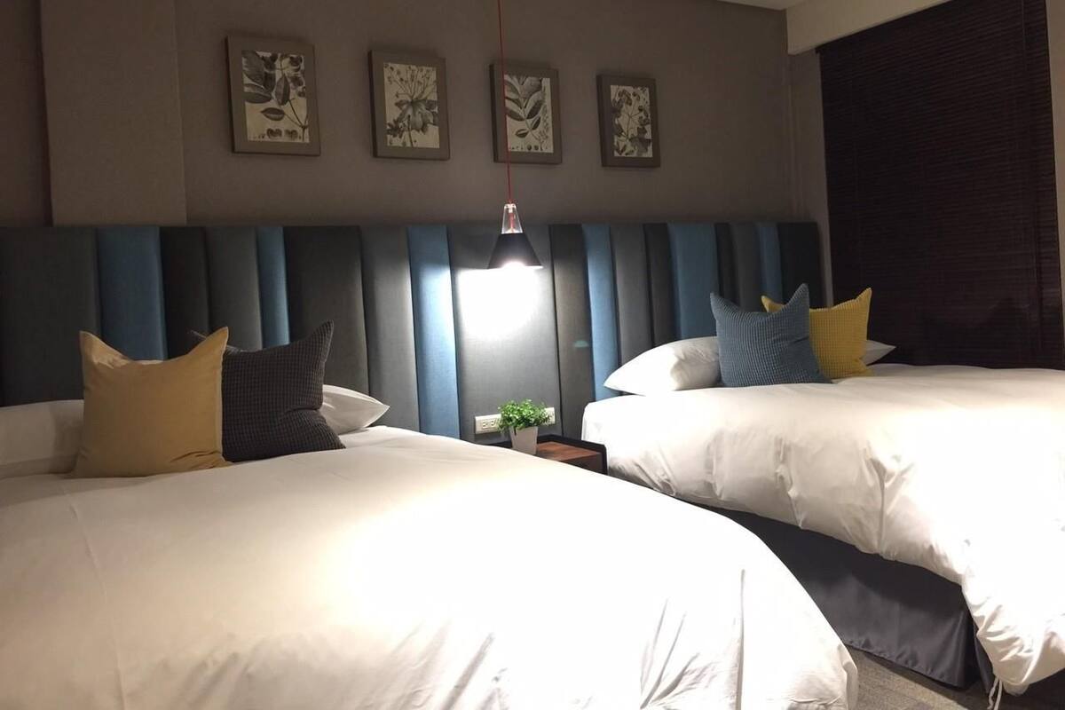 A hotel room with two beds, a nightstand, a wall-mounted lamp, and framed floral artwork.