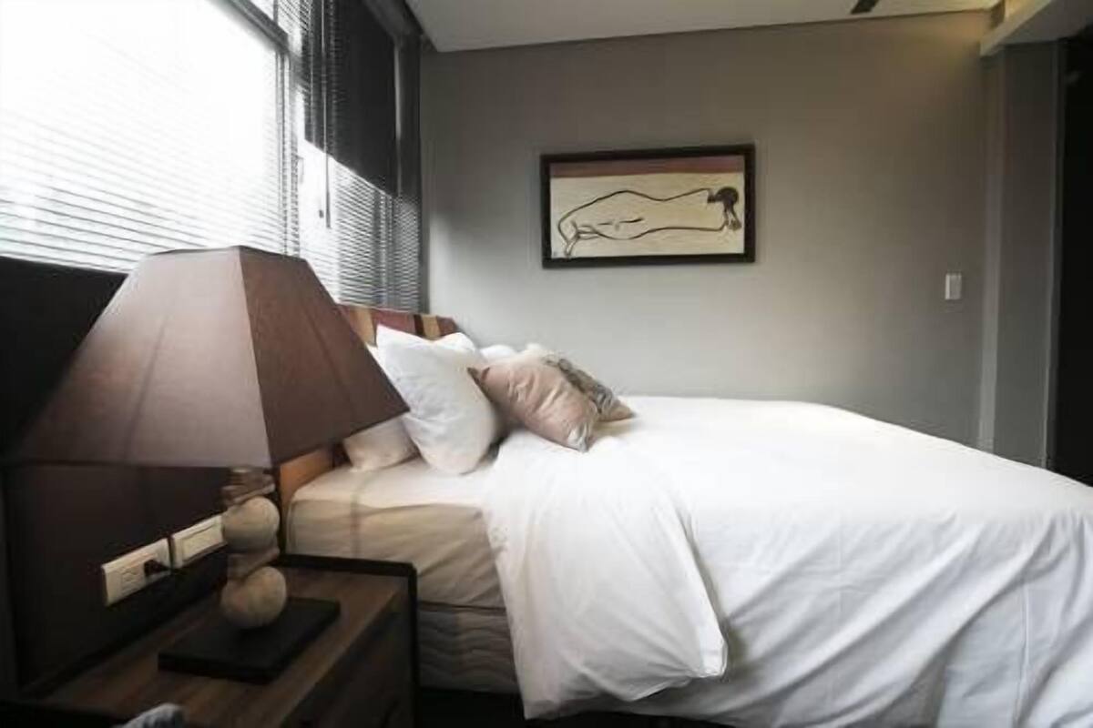 A neatly made bed with white linens and a few pillows, a bedside lamp, and a framed artwork on the wall.