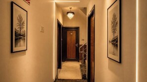 Un pasillo con cuadros enmarcados, un cartel de 'No Fumar', una araña y una puerta de madera al final.