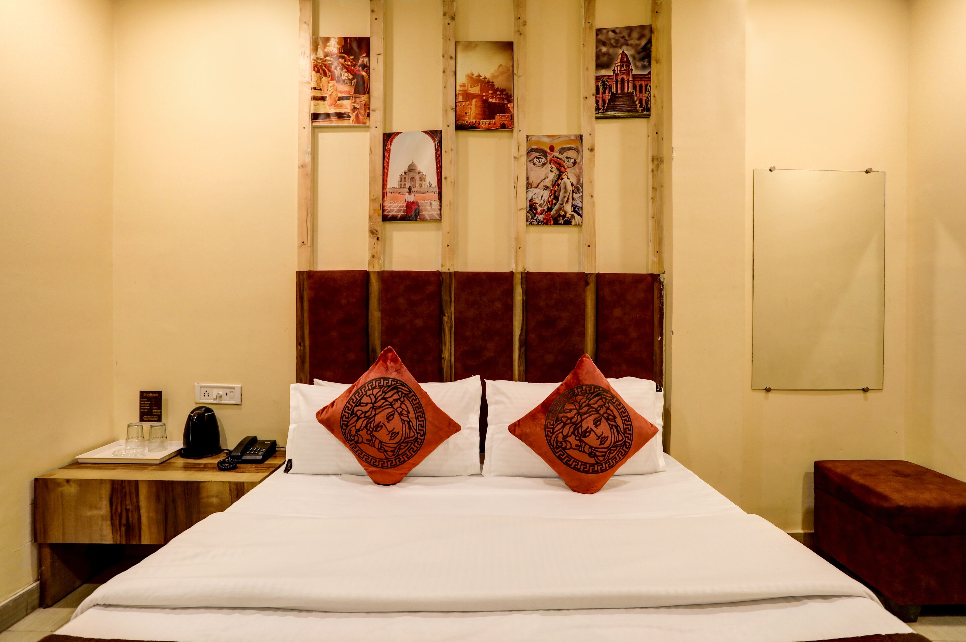 A hotel room with a large bed, two decorative pillows, a wooden nightstand with a phone and glasses, and a red bench.