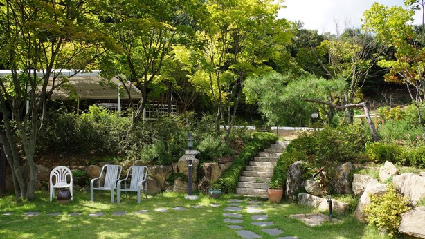 A garden with stone steps leading to a house, surrounded by greenery and two white chairs.