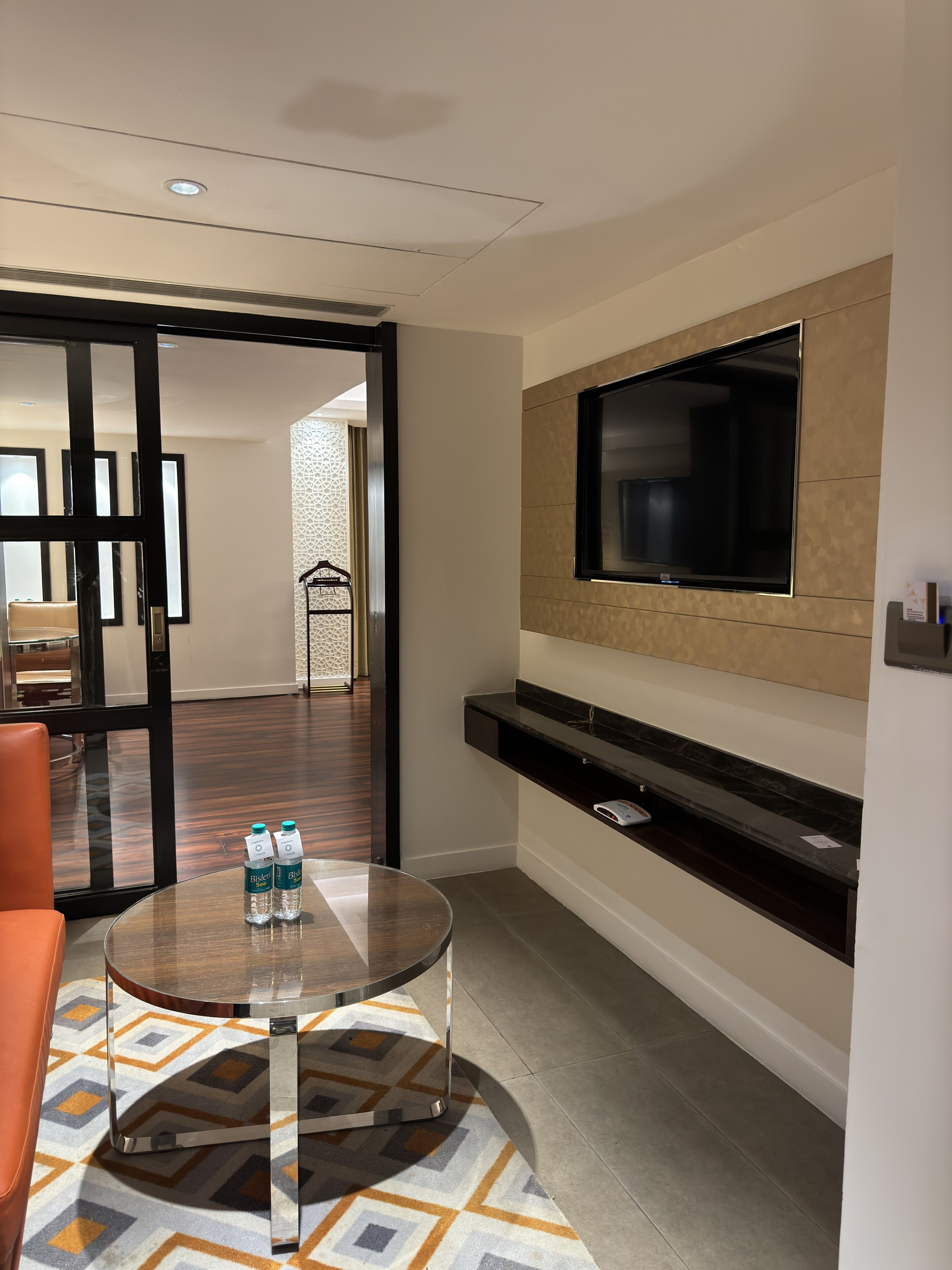 A hotel room with a glass table, a sofa, a TV mounted on the wall, and a patterned rug.