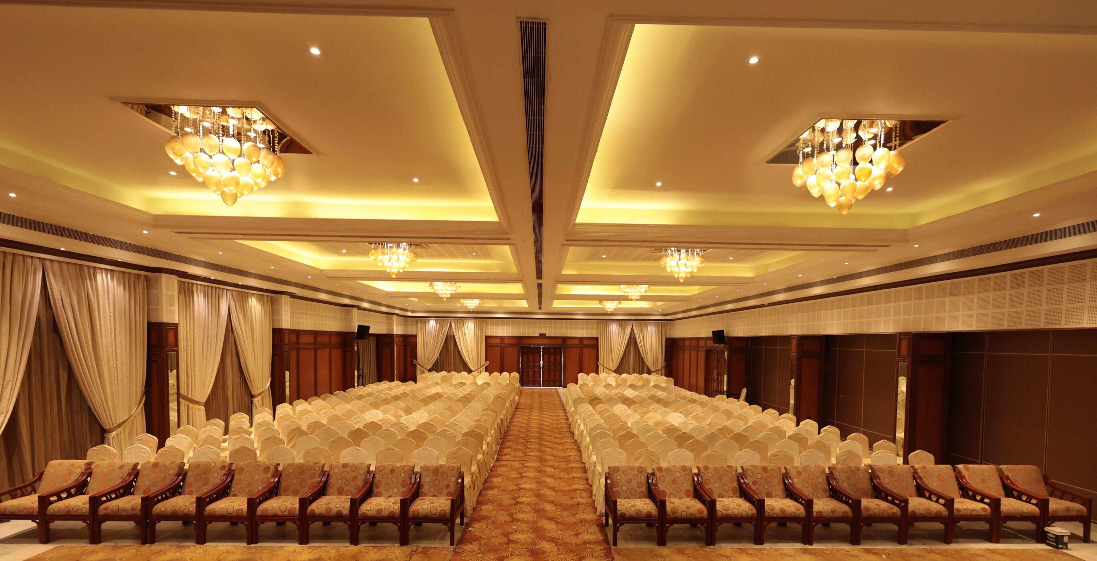 A conference room with rows of chairs, a carpeted floor, chandeliers, and curtains.
