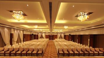 A conference room with rows of chairs, a carpeted floor, chandeliers, and curtains.
