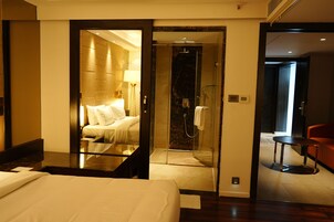 Habitación de hotel con cama, zona de estar con mesa y sillas, cabina de ducha y una silla roja en la esquina.