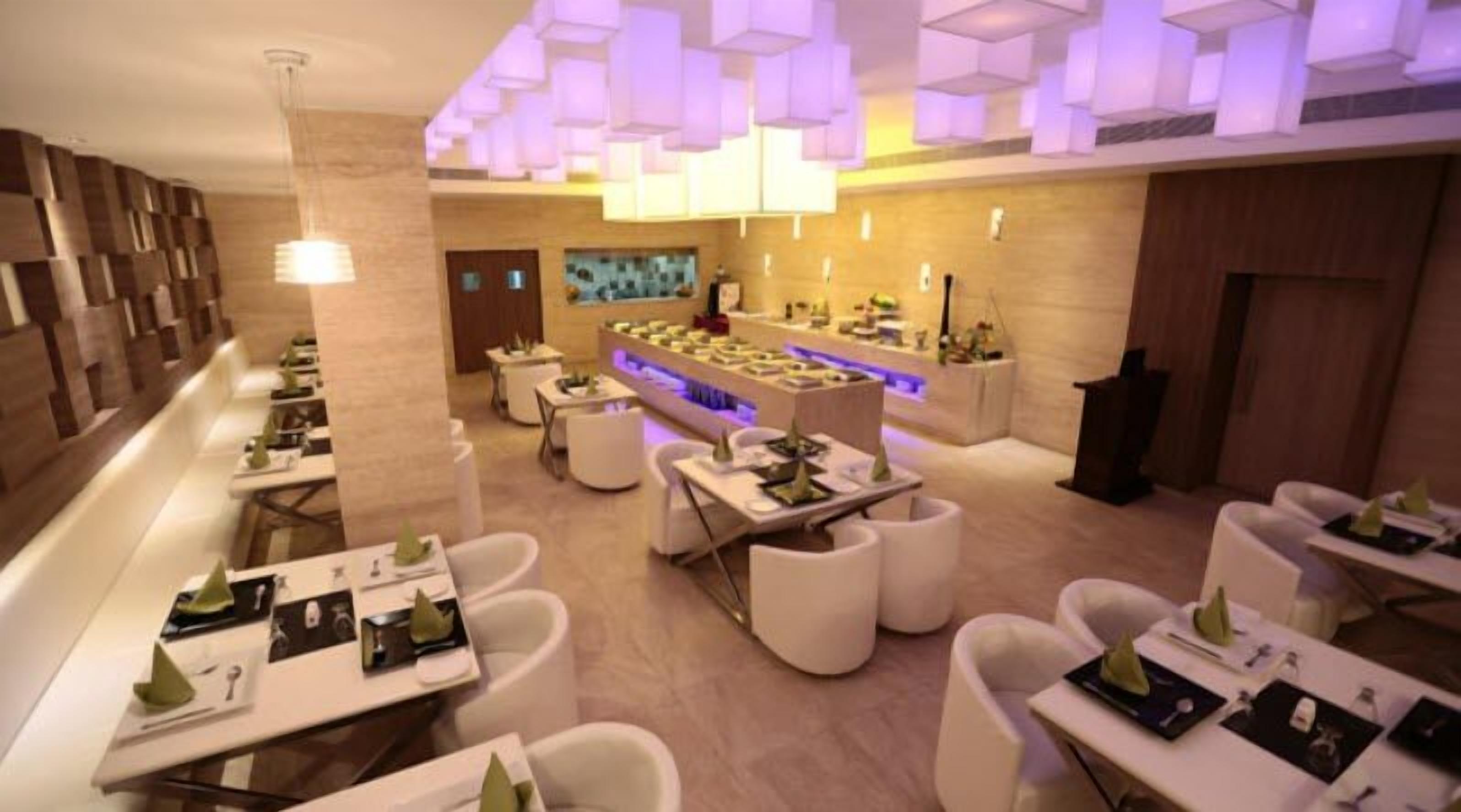 A modern restaurant with white tables and chairs, a central bar area, and a ceiling with geometric light fixtures.