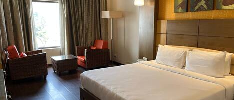A hotel room with a large bed, a sitting area with a chair and table, a lamp, and artwork on the wall.