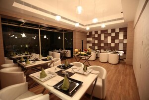 Un comedor moderno con mesas dispuestas para el servicio, un muro a cuadros y lámparas colgantes.