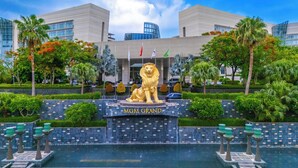 Uma entrada imponente com uma estátua dourada de leão, piscina e fachada de edifício moderna com bandeiras.