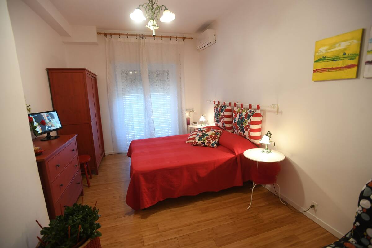 A bedroom with a red bed, a bedside table with a lamp, a wooden wardrobe, a TV on a desk, and a window with curtains.