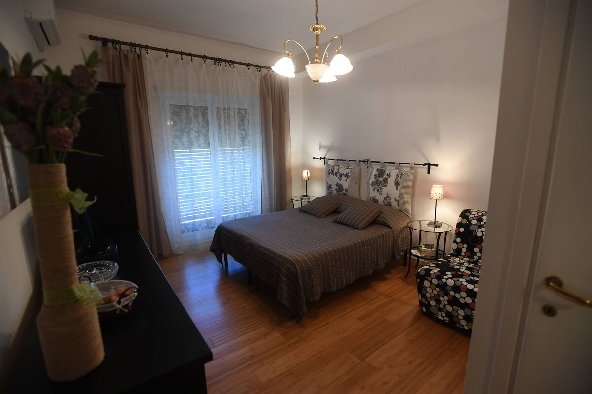 A bedroom with a bed, bedside lamps, a small table, a chair, and a vase with flowers.