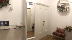 A hallway with a glass shelf displaying books, a door with a sign, a mirror, and a chair with a floral arrangement. - A Casa di Enza (Rome)