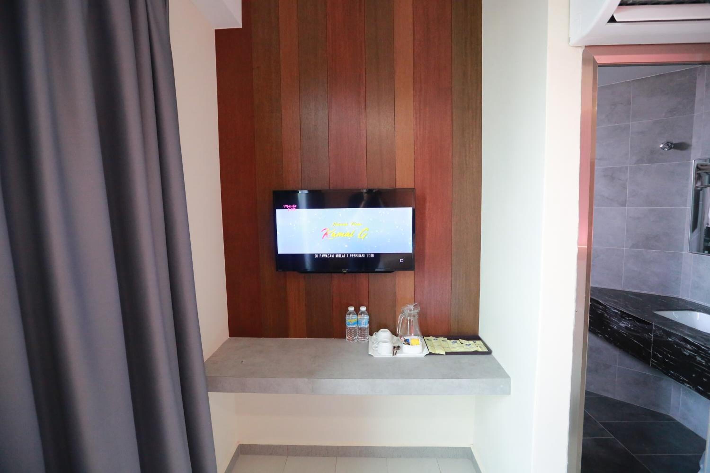 A TV mounted on a wooden panel wall, a small shelf with bottled water, a coffee maker, and a tray with papers.