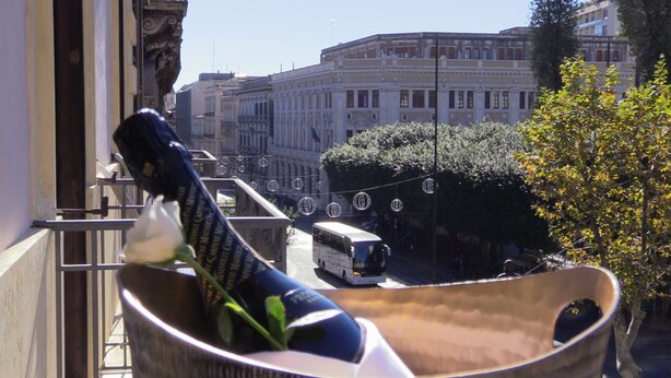 A balcony with a wicker chair, a white rose, and a view of a city street with buildings and a bus. - Moonlight Hotel & Suites (Catania)