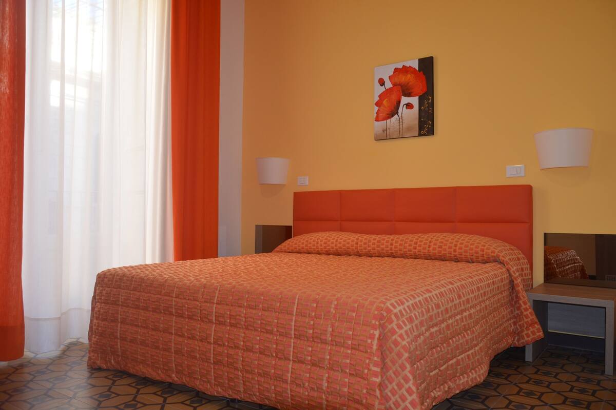 A hotel room with a large bed, two bedside tables, a wall-mounted lamp, and a painting of flowers.
