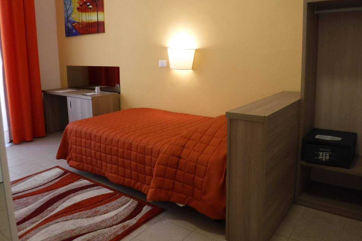 A hotel room with a bed, a desk, a lamp, a painting, and a wardrobe.