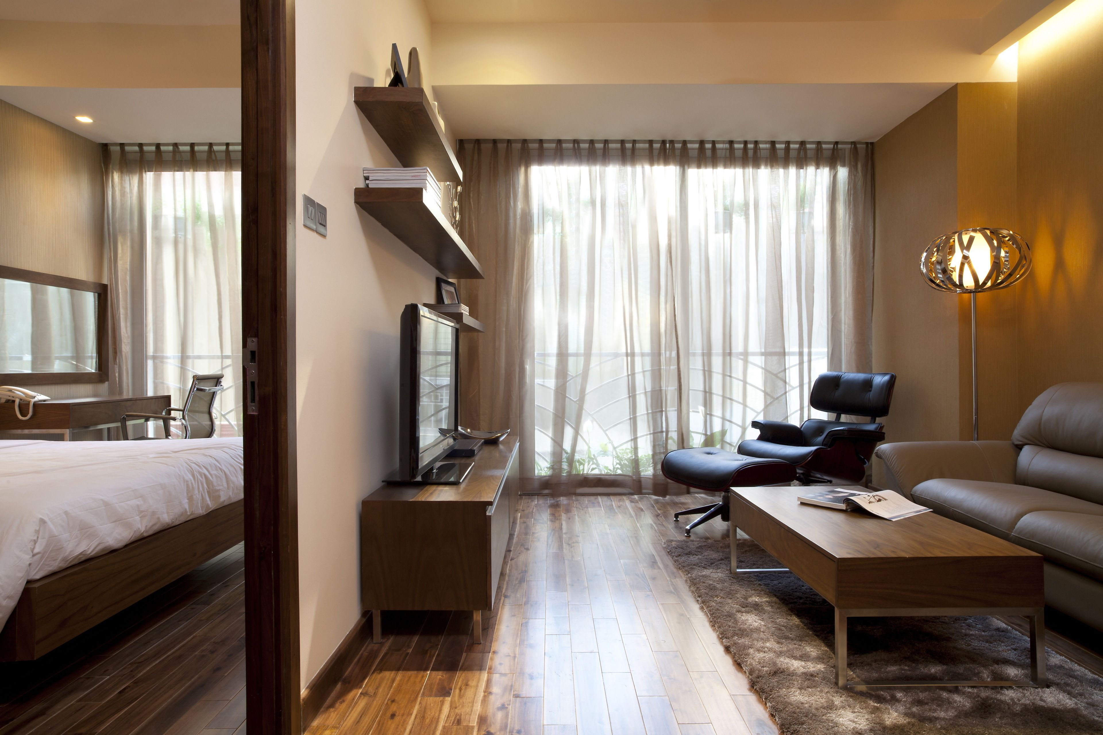 A modern hotel room with a wooden bed, a desk, a leather sofa, and a large window with sheer curtains.