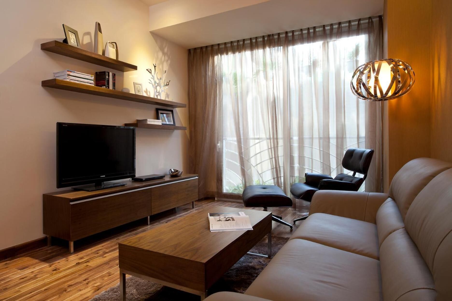 A modern living room with a flat-screen TV, wooden shelves, a leather sofa, and a large window with sheer curtains.