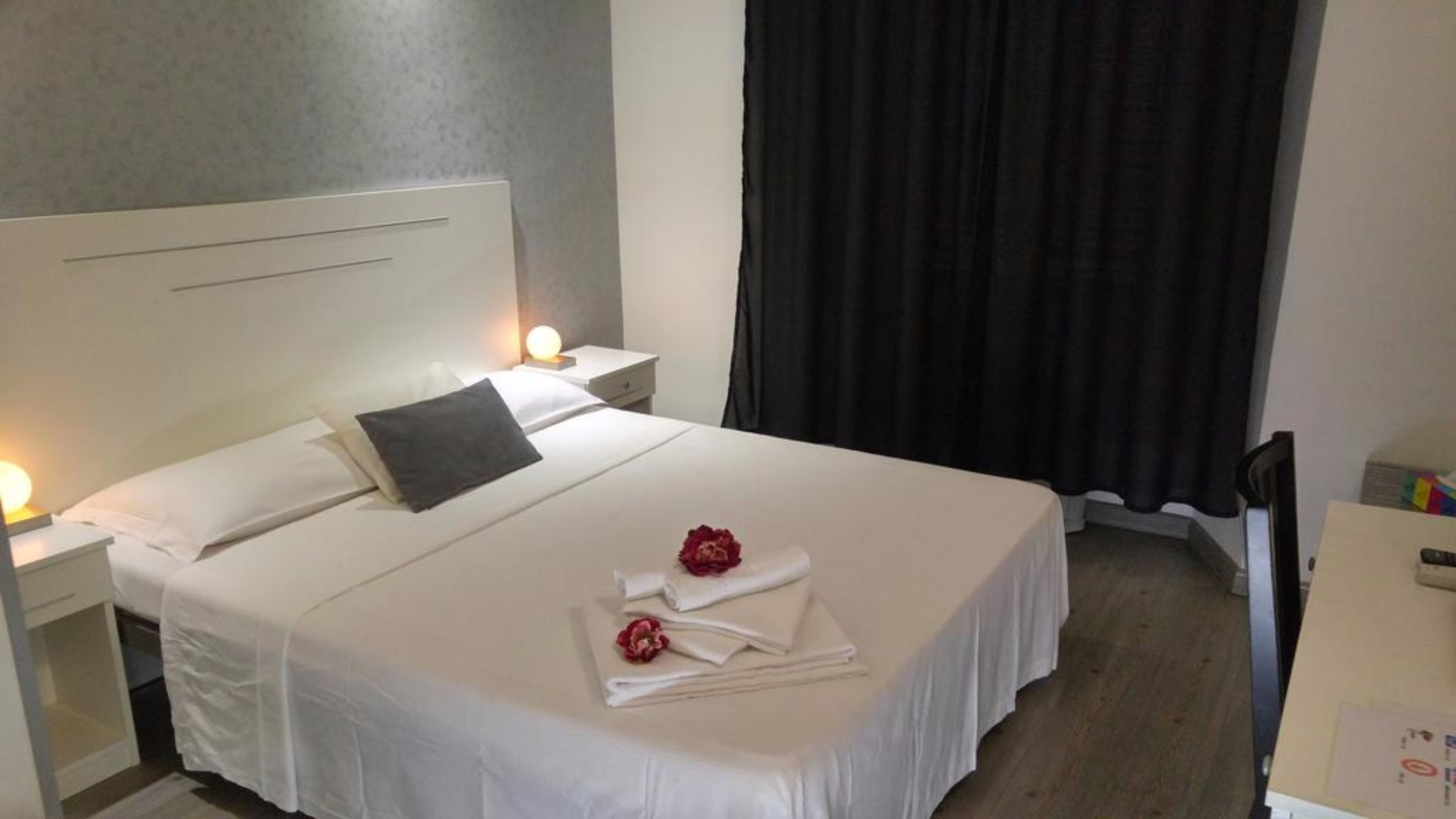 A neatly made bed with white linens, a dark pillow, and two red rose bouquets on the bed. There are two bedside tables with lamps, a dark curtain, and a wooden floor.