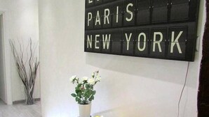 Un interior moderno con un letrero negro en la pared que menciona 'LONDON PARIS NEW YORK'. Hay una mesa de vidrio con un frutero y una planta en maceta.