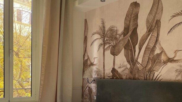 Habitación de hotel con cama, ropa de cama blanca, cabecera, ventana con cortinas y un mural de hojas tropicales.