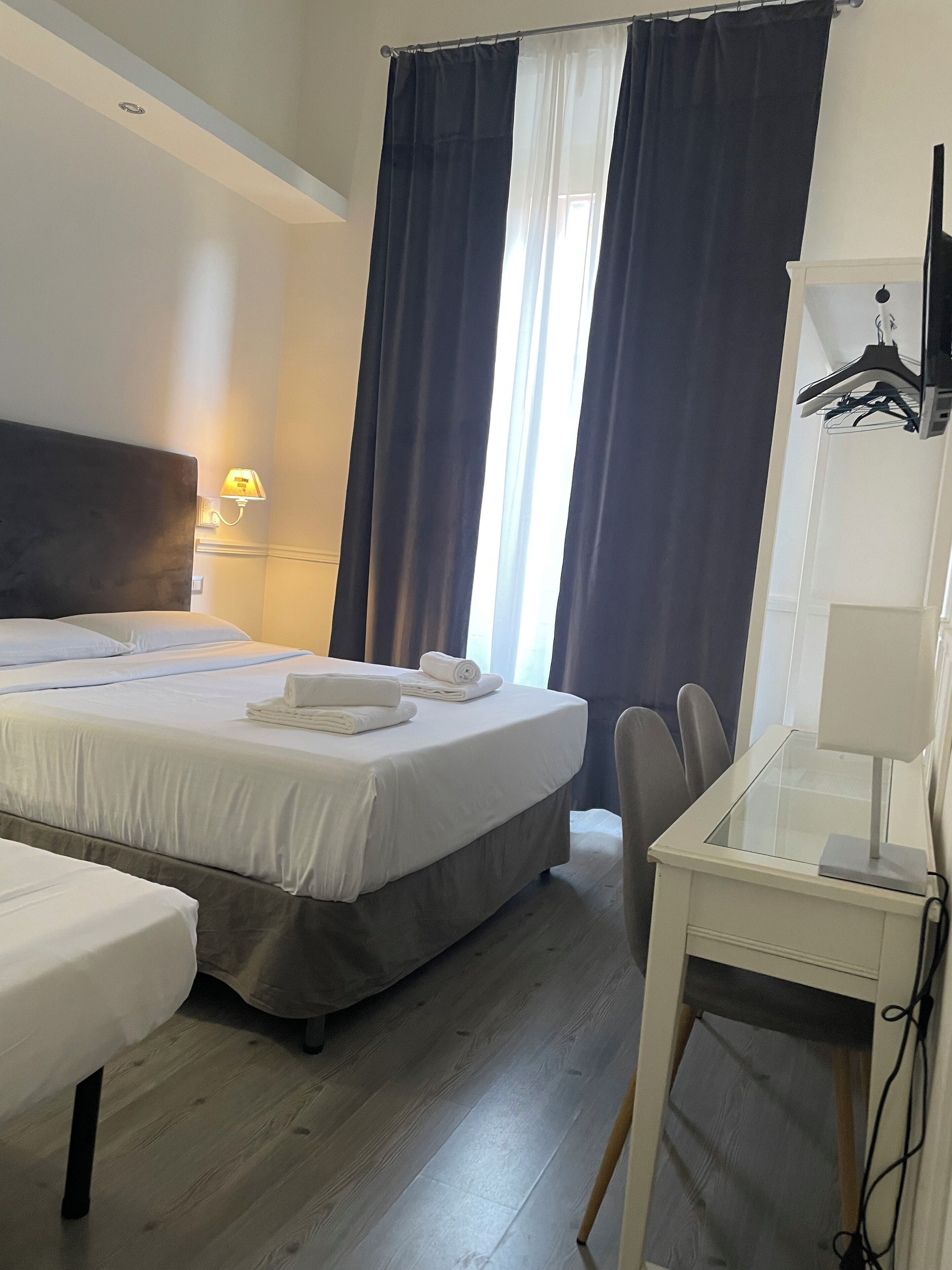 Une chambre d’hôtel avec un lit, un bureau et une chaise. Il y a une fenêtre avec des rideaux, une lampe et un porte-manteau.