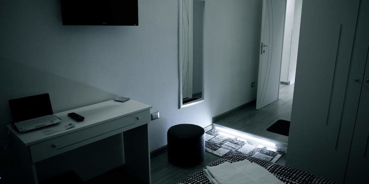 A small, dimly lit room with a desk, a chair, a laptop, a remote control, a trash bin, and a window with a view of a door and a hallway.