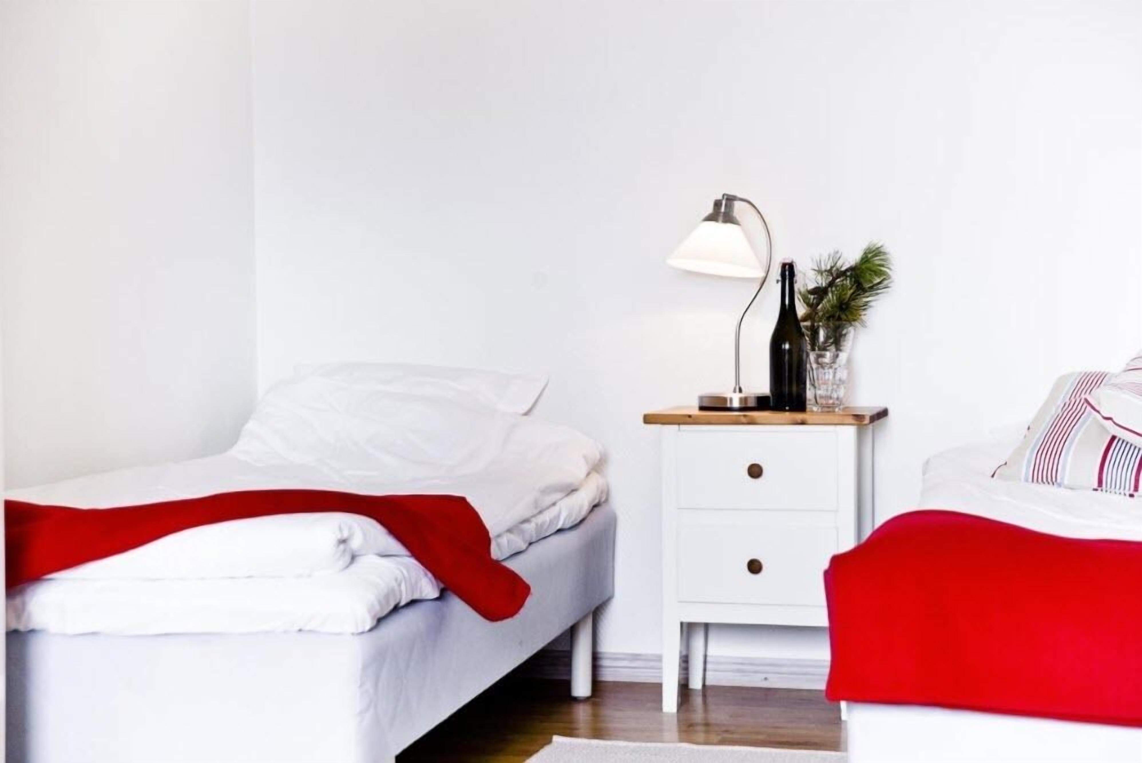 A neatly made bed with white bedding and a red throw blanket, a bedside table with a lamp and a small plant, and a white nightstand with drawers.
