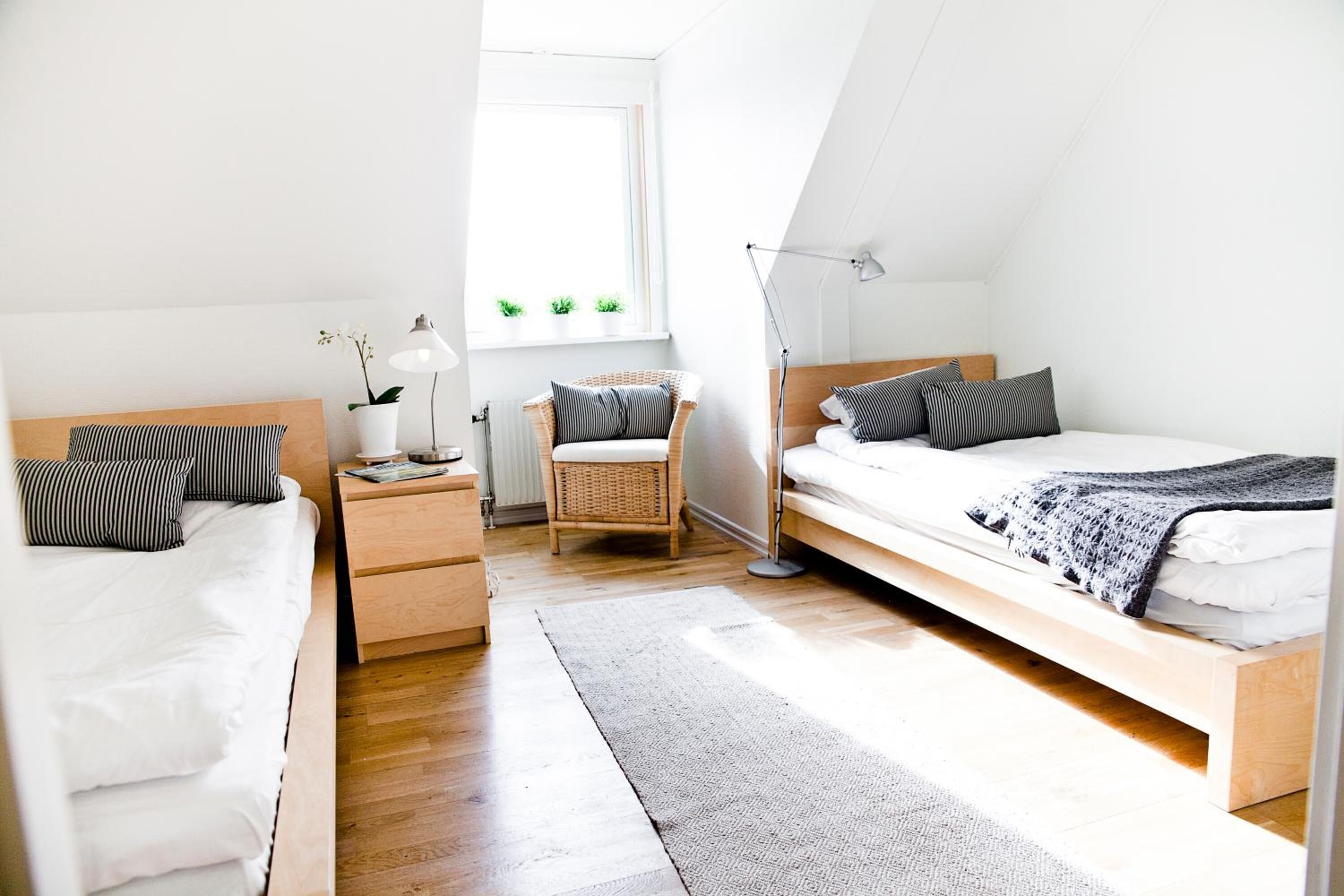 Un dormitorio moderno y bien iluminado con dos camas, una mesita de noche, una silla y una ventana con plantas.