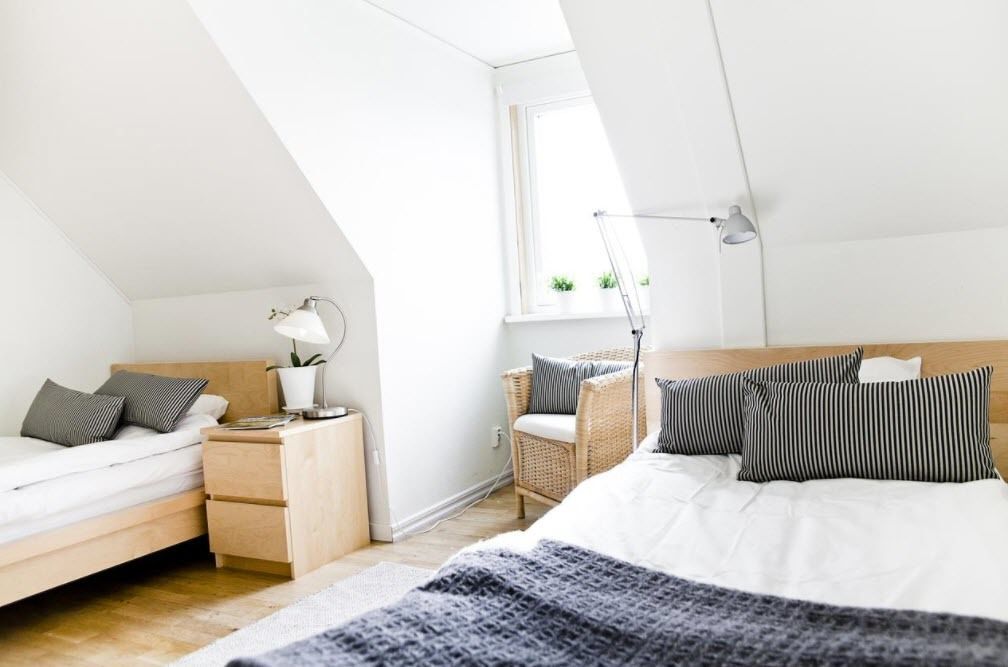 A modern bedroom with a sloped ceiling, a bed with striped pillows, a bedside table with a lamp, a wicker chair, and a window with plants.