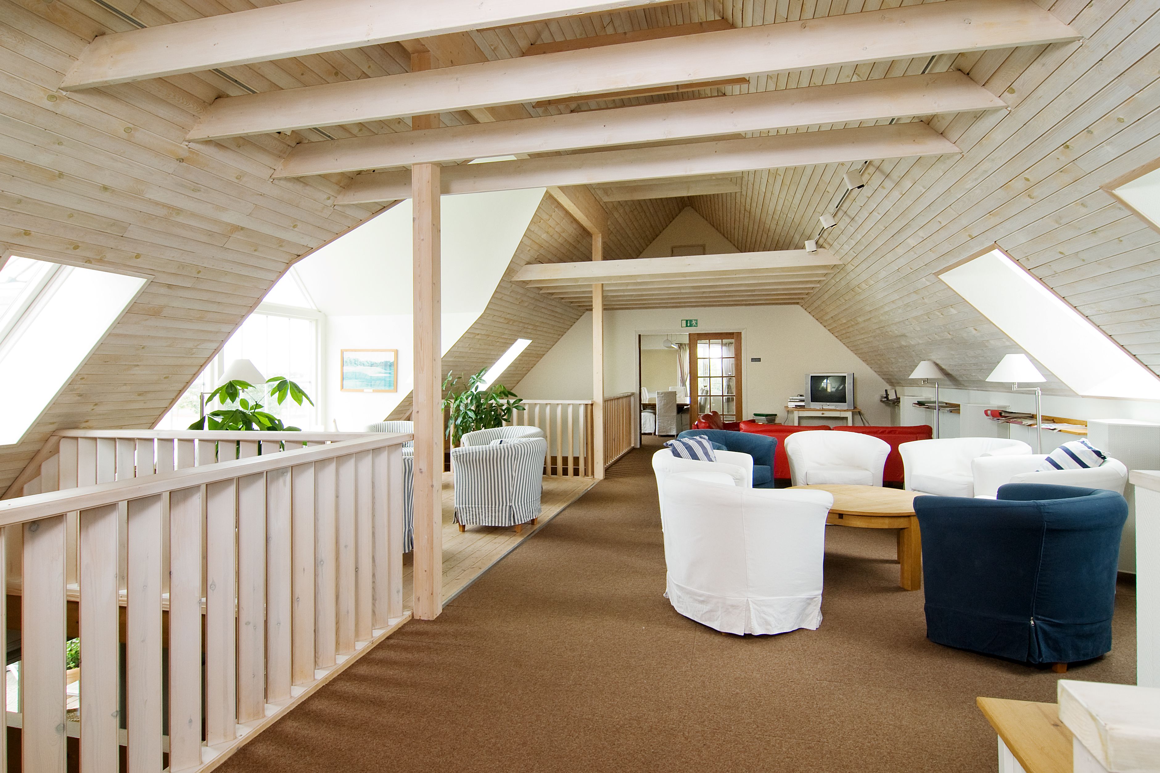 Een ruime loft-achtige kamer met hoge plafonds, houten balken en een schuin dak. Er is een gezellige zithoek met wit en blauw gestoffeerde stoelen, een kleine tafel en een pot met een plant.