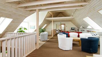 Een ruime loft-achtige kamer met hoge plafonds, houten balken en een schuin dak. Er is een gezellige zithoek met wit en blauw gestoffeerde stoelen, een kleine tafel en een pot met een plant.