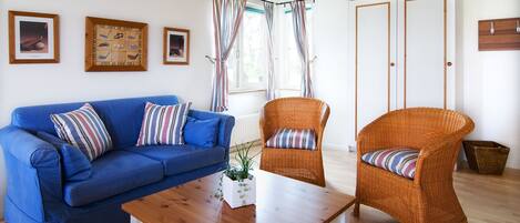 Uma sala de estar com um sofá azul, cadeiras de vime, uma mesa de centro de madeira e quadros nas paredes.