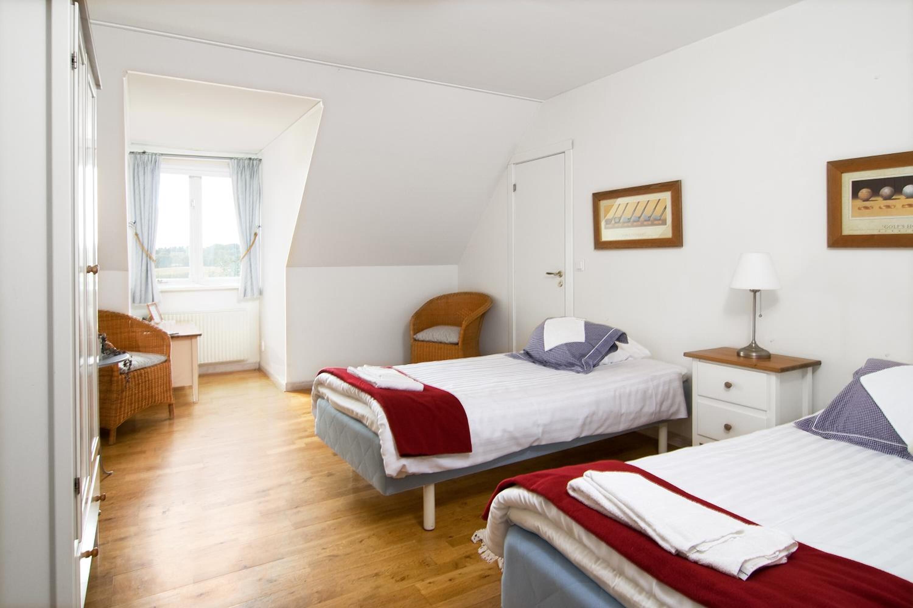 A bedroom with two beds, a window with curtains, a chair, a bedside table, and a lamp.