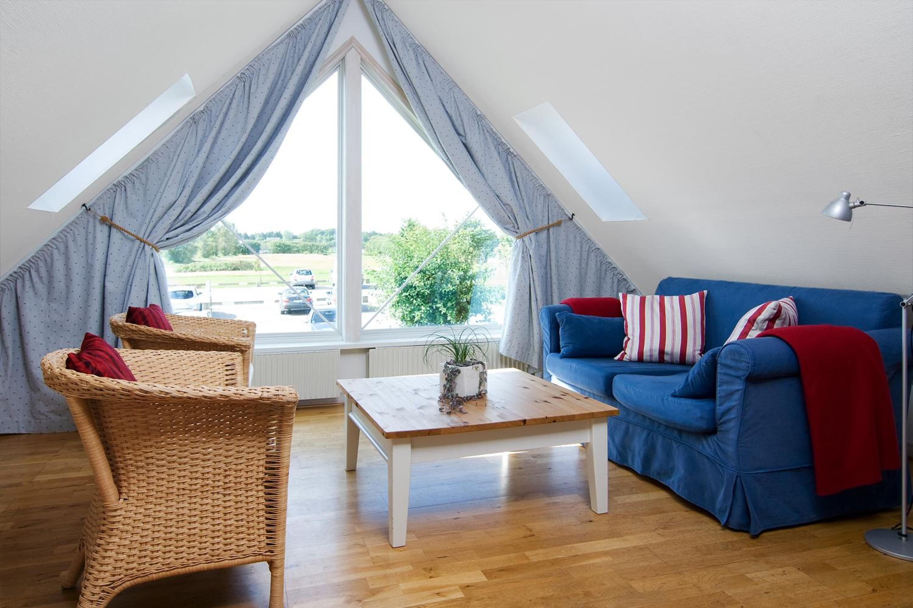 Un salon chaleureux avec un canapé bleu, deux fauteuils en osier, une table basse en bois et une grande fenêtre avec des rideaux.