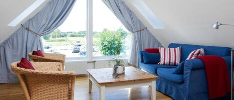 Un salon chaleureux avec un canapé bleu, deux fauteuils en osier, une table basse en bois et une grande fenêtre avec des rideaux.