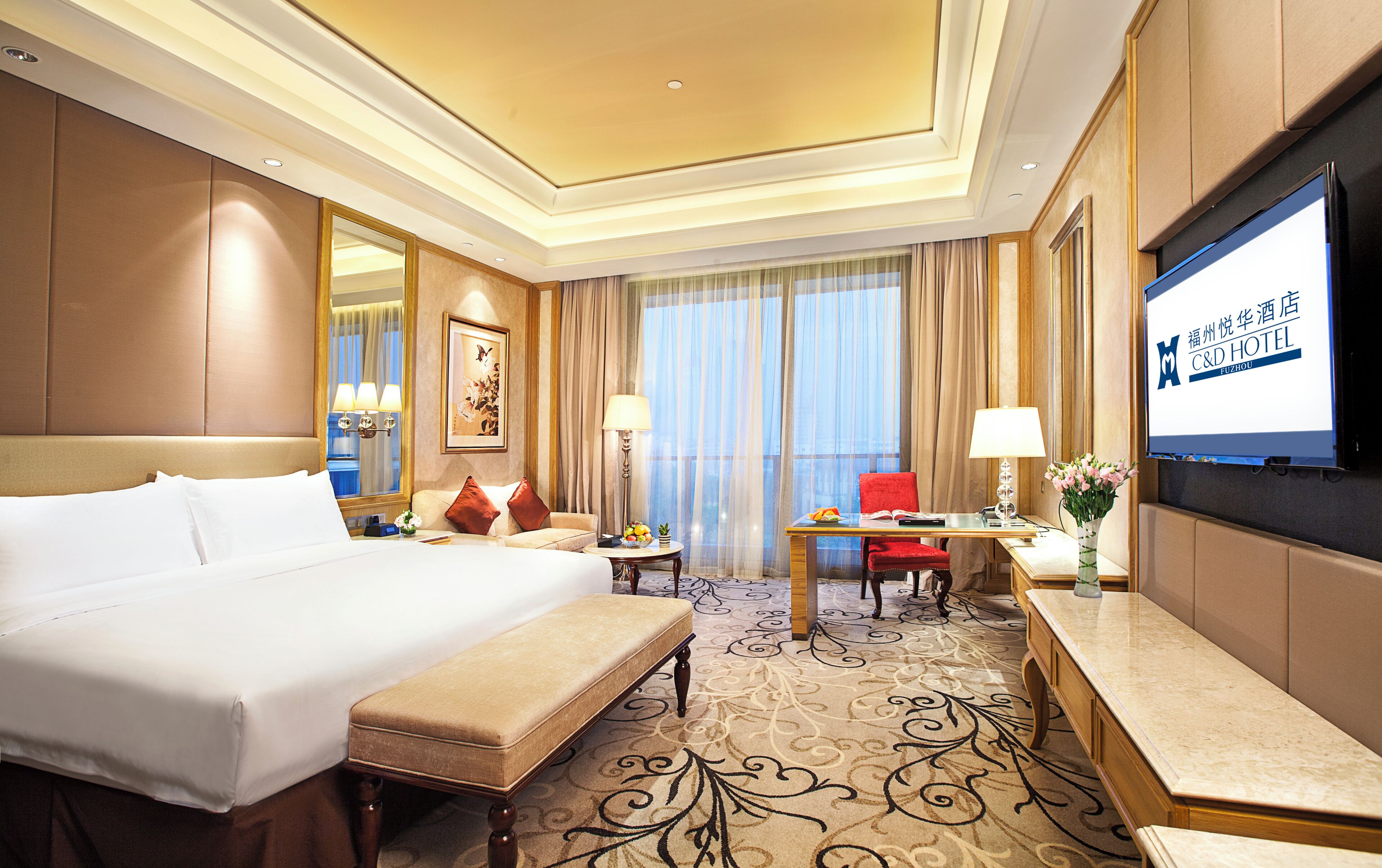 A hotel room with a large bed, a desk, a chair, and a TV screen displaying hotel information.