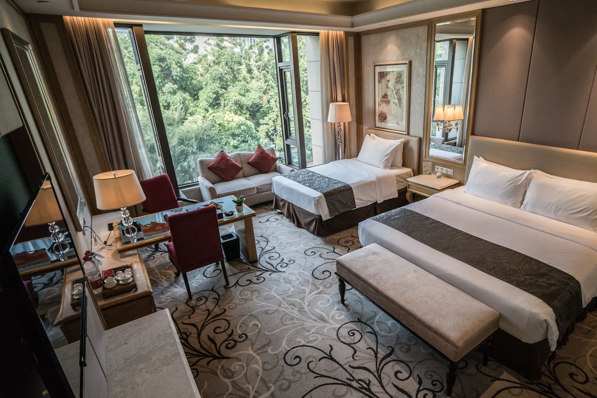 A hotel room with two beds, a desk, a chair, and a large window offering a view of trees.