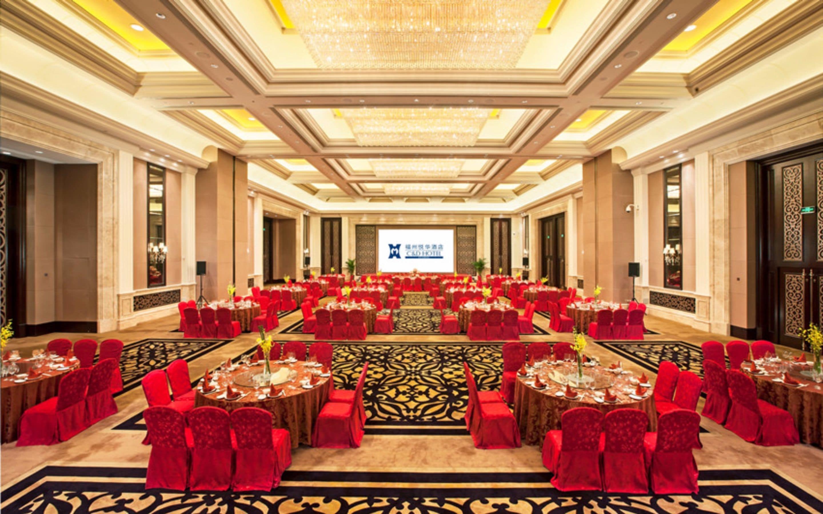A spacious banquet hall with round tables and red chairs, a chandelier, and a large screen displaying the hotel name.
