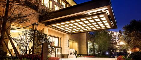 A well-lit hotel entrance with a covered walkway, a statue, and landscaped gardens.