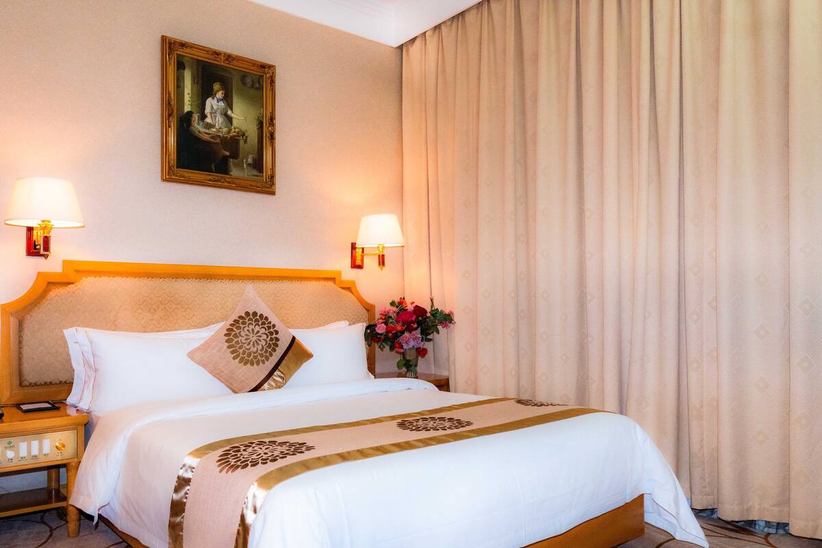 Una habitación de hotel con una cama grande, dos lámparas de noche, una mesita de noche, un cuadro en la pared y una cortina.