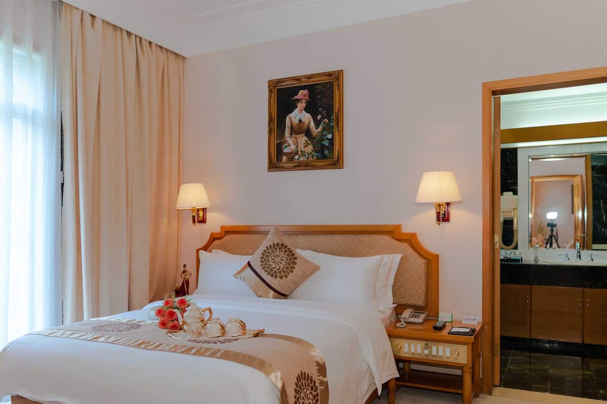 Una habitación de hotel con una cama grande, dos lámparas de noche, un cuadro sobre la cama, un espejo y una mesita de noche con un teléfono y una lámpara.
