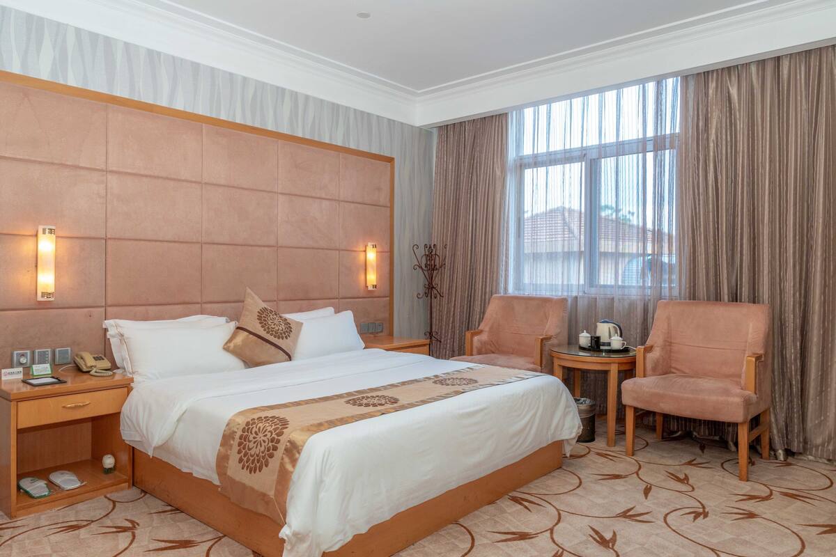 Habitación de hotel con una cama grande, dos mesitas de noche, una silla, una mesita pequeña con una tetera y una ventana con cortinas.