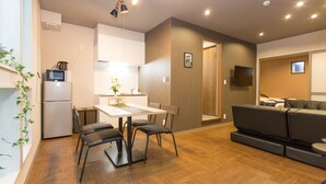 A modern living room with a sofa, a dining table with chairs, a kitchen area with a refrigerator, and a TV mounted on the wall. - Group STAY CLASSE 02 (Osaka)