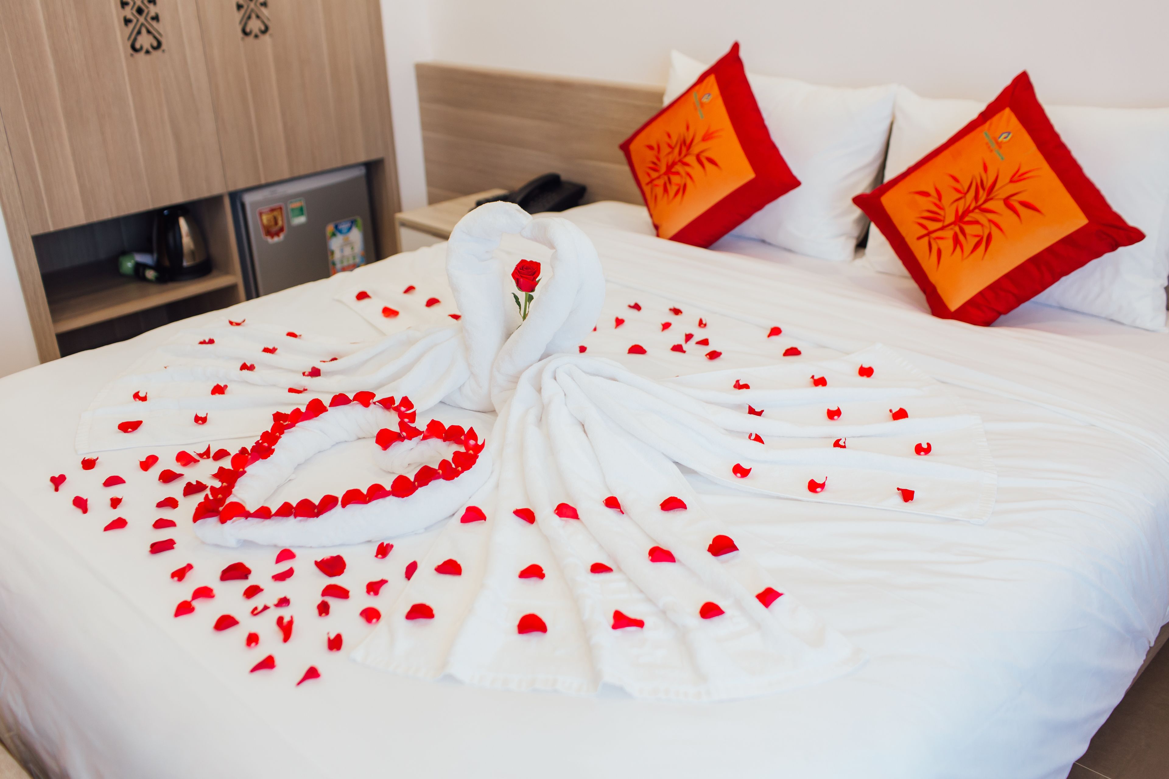 A neatly made bed with a swan and rose decorations, a phone on the nightstand, and a minibar with a kettle and a bottle.