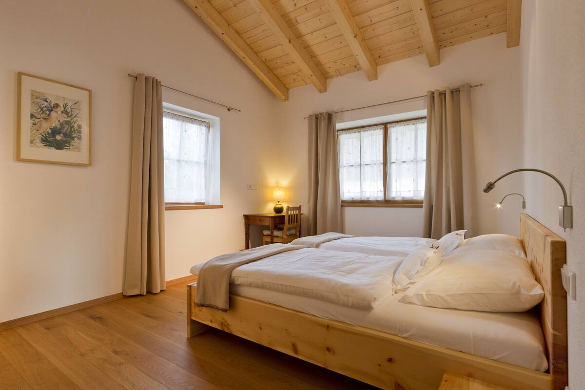 Et soveværelse med en træseng, to vinduer med gardiner, et skrivebord med lampe og et billede i ramme på væggen.
