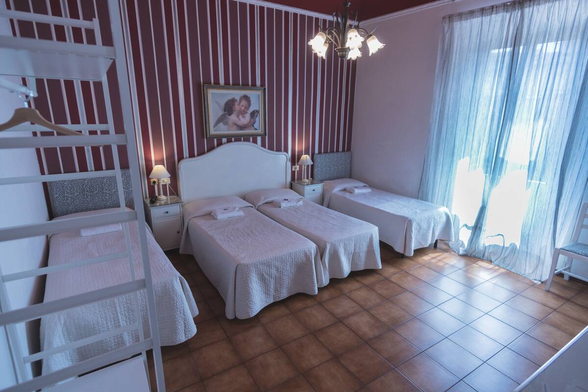 A hotel room with two single beds, a wooden ladder, a nightstand with a lamp, a framed picture on the wall, and a large window with sheer curtains.